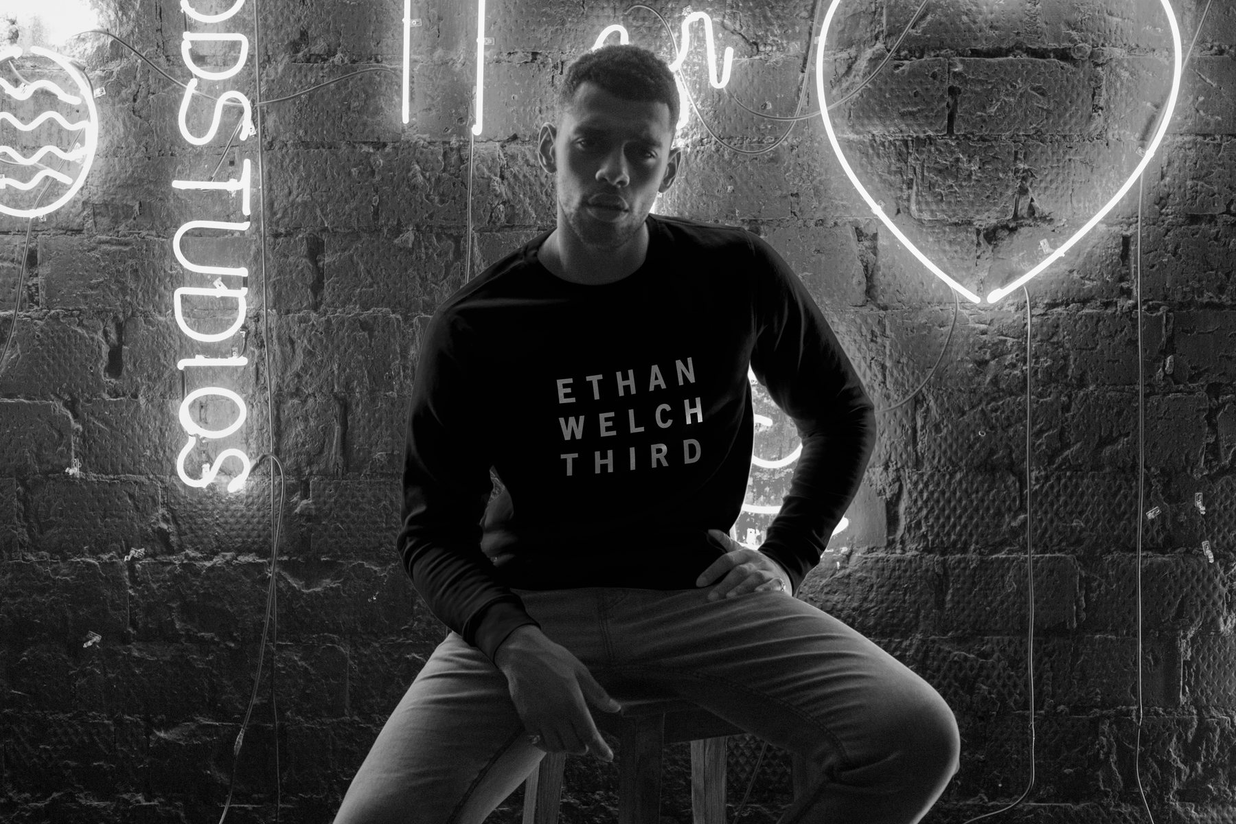 A black sweatshirt with the stacked wordmark, worn in front of a neon-lit brick wall
