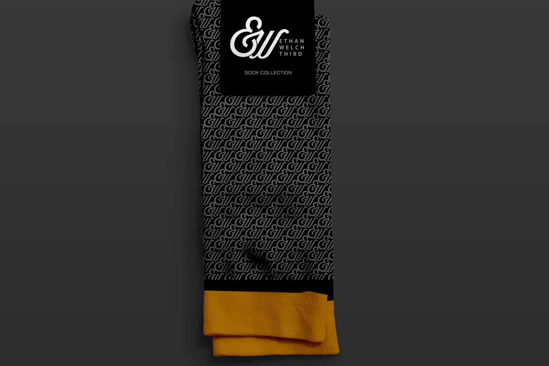 A folded black sock with a mustard cuff and the monogram pattern, with its black hang tag