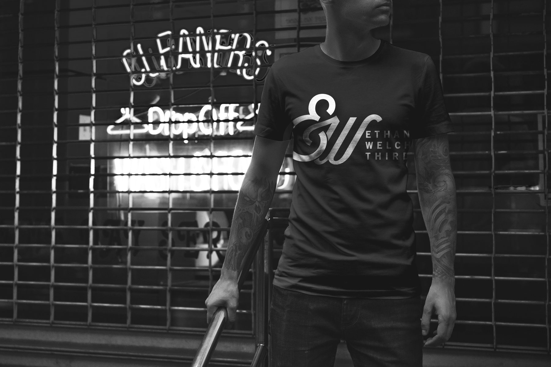 A black tee with the monogram and wordmark, worn in front of a shuttered storefront
